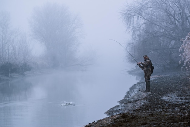 Un pescador pescando con una caña giratoria en una orilla del río en un invierno brumoso y brumoso pescando presas pescando