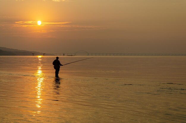 Un pescador se para sobre una roca en el mar al amanecer.