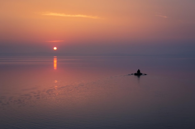 Pescador solitario en un pequeño bote flota en un lago tranquilo contra el colorido amanecer