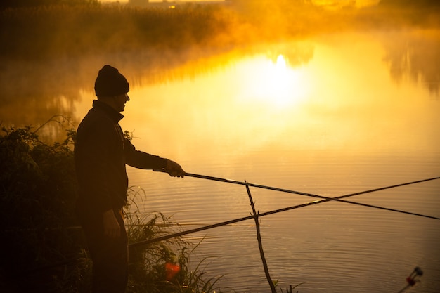 Pescador solitario pescando en el lago brumoso temprano en la mañana, justo después del amanecer dorado.