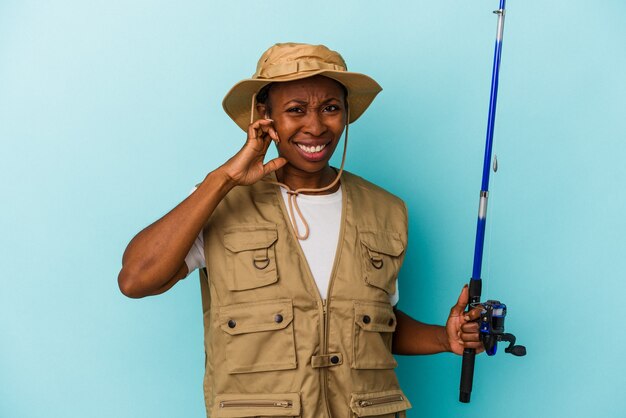 Pescadora afroamericana joven sosteniendo la barra aislada sobre fondo azul cubriendo las orejas con las manos.