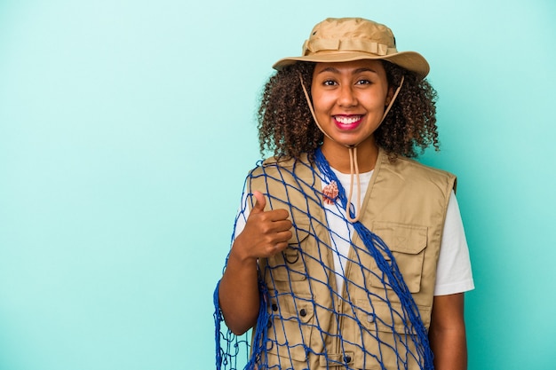 Pescadora afroamericana joven sosteniendo net aislado sobre fondo azul sonriendo y levantando el pulgar hacia arriba