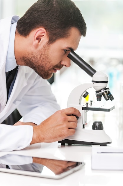 Pesquisa e desenvolvimento. Vista lateral de um jovem cientista de uniforme branco usando um microscópio enquanto está sentado em seu local de trabalho