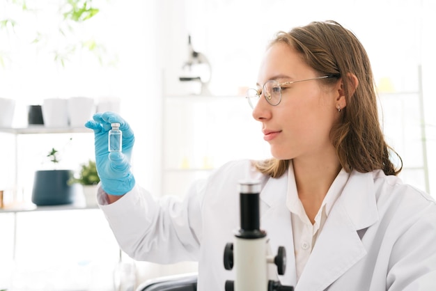 Pesquisadora cientista caucasiana olhando e segurando uma pequena garrafa de vidro médio para análise química de líquidos no laboratório Cientista trabalhando com equipamentos químicos no laboratório