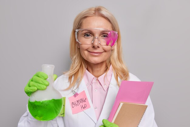 Pesquisadora satisfeita usando óculos de proteção jaleco branco médico segura frasco de vidro com caderno de líquido verde para escrever notas realiza experimento científico isolado em cinza