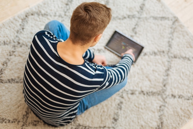 Pesquisando na net. Jovem estiloso de cabelos louros, bem constituído, vestindo roupas casuais e sentado no chão usando seu tablet