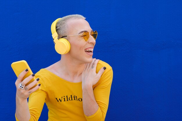 Pessoa alegre não binária que gosta de ouvir música com fones de ouvido sem fio e um telefone celular, ao ar livre, contra uma parede azul.