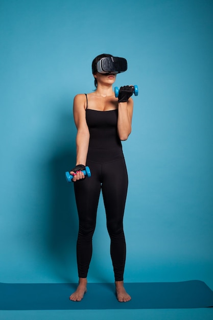 Pessoa ativa usando músculos do braço de treinamento de fone de ouvido de realidade virtual usando halteres trabalhando na resistência do corpo. Personal trainer em roupas esportivas em pé no tapete de ioga praticando exercícios de fitness