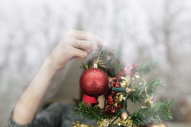 Pessoa através de uma janela pendurando decorações de natal na árvore com uma bugiganga vermelha na mão e reflexos no vidro.