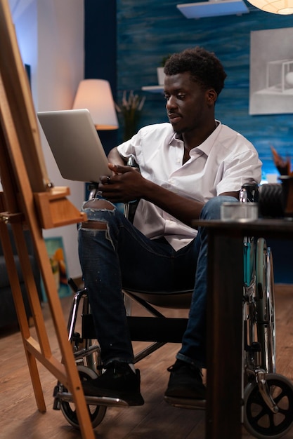 Pessoa com habilidades artísticas e deficiências fazendo cursos de desenho online com laptop. Usuário de cadeira de rodas afro-americano em busca de inspiração em sites de arte para criar esboços de obras-primas no atelie