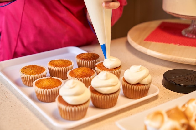 Pessoa de colheita irreconhecível decorando cupcakes usando saco de tubulação de pé no balcão da cozinha com outras sobremesas nele