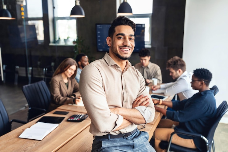 Foto pessoa de sucesso empresário árabe confiante, apoiando-se na mesa no escritório, posando com os braços cruzados e sorrindo para a câmera
