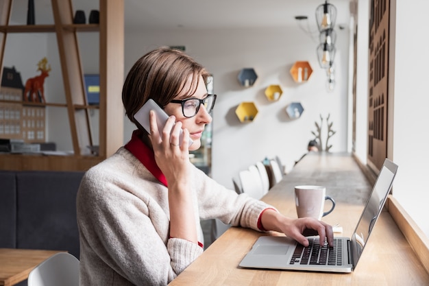 Pessoa do sexo feminino falando ao telefone e usando o laptop na hora do almoço em um café. mulher de negócios de conteúdo trabalhando com tecnologia em um local público