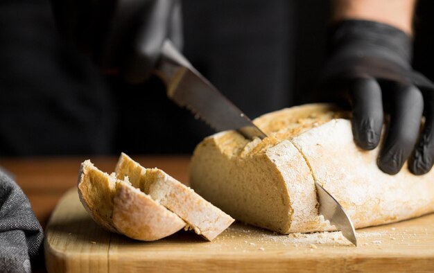 Pessoa em luvas de cozinha preta cortando pão fresco deitado em uma tábua de madeira