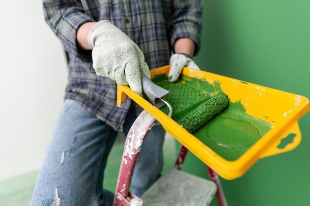 Pessoa em uma escada de escada em luvas de trabalho segurando uma caldeira de pintura amarela com um rolo de pintura de tamanho médio com pintura verde na frente da parede verde