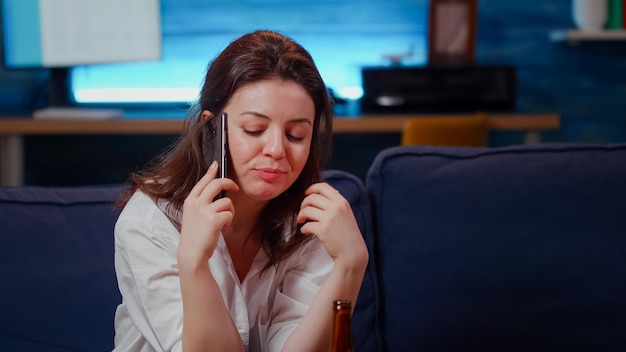 Pessoa falando ao telefone com um amigo na sala de estar enquanto come batatas fritas e relaxa. Mulher caucasiana se divertindo conversando no smartphone, sentada no sofá com lanche depois do trabalho