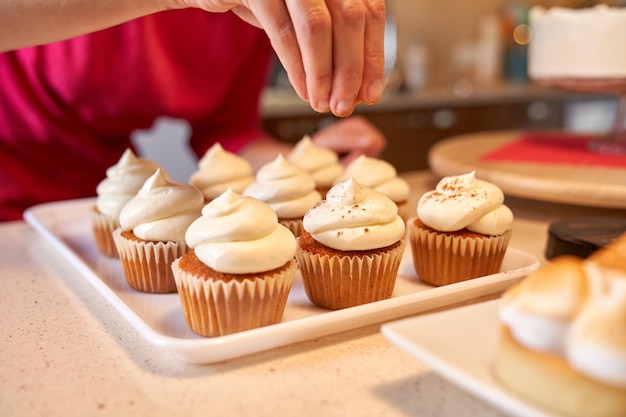 Foto pessoa irreconhecível de pé no balcão da cozinha com várias sobremesas e pulverizando canela em cupcakes decorados com creme de manteiga