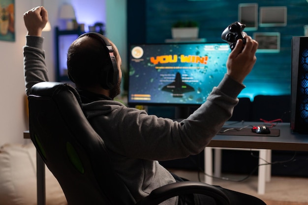 Pessoa jogando videogame com controlador e ganhando na frente do computador. Jogador usando joystick e fones de ouvido para jogar jogos online. Homem usando fone de ouvido e comemorando a vitória.