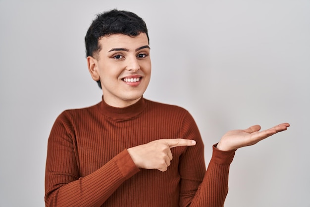 Pessoa não binária usando maquiagem em pé sobre um fundo isolado espantado e sorrindo para a câmera enquanto apresenta com a mão e aponta com o dedo.