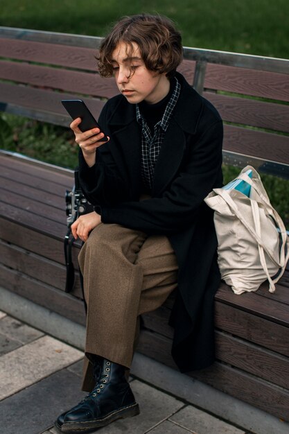 Pessoa não binária verificando o telefone enquanto está sentada em um banco