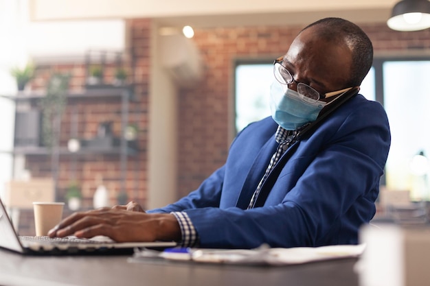 Pessoa no trabalho usando smartphone para ter uma ligação com o gerente durante a pandemia de covid 19, trabalhando com o laptop para planejar o projeto. empreendedor usando máscara facial e falando no celular.