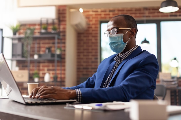 Pessoa olhando para a tela do laptop para planejar o projeto de negócios durante a pandemia de coronavírus. Empreendedor com máscara facial usando o computador para trabalhar na estratégia de marketing no escritório de inicialização.