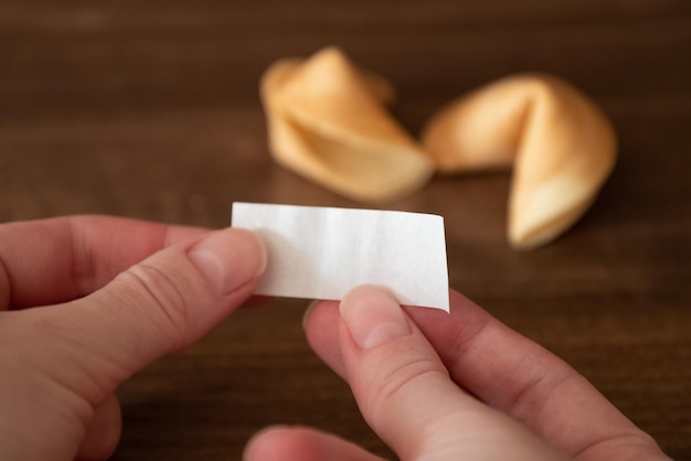 Pessoa segura nas mãos o papel em branco do biscoito da sorte contra dois biscoitos deitados na mesa