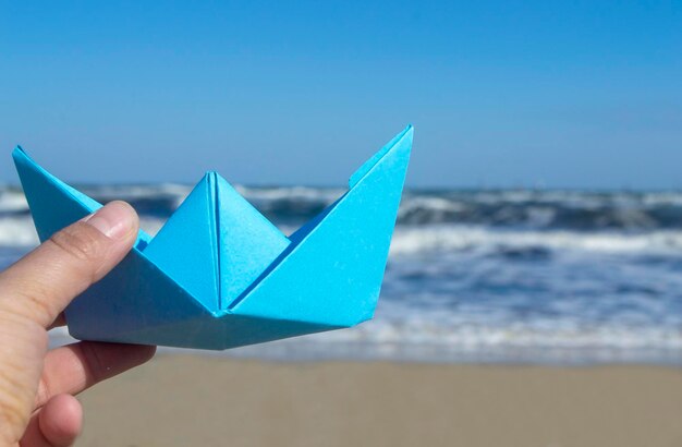 Foto pessoa segurando o barco de papel azul na mão no fundo do céu azul das ondas do mar