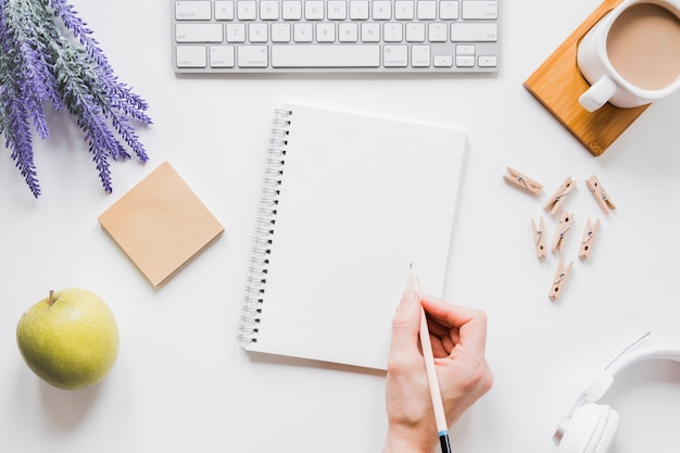 Foto pessoa sem rosto, escrevendo no caderno na mesa branca com uma xícara de café e teclado