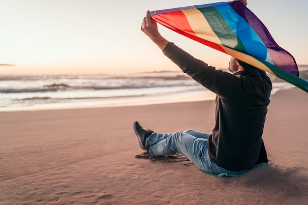 pessoa sentada na praia e segurando a bandeira do arco-íris acima da cabeça vista traseira LGTBQ