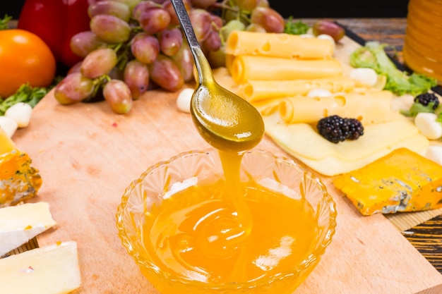 Pessoa servindo mel dourado delicioso de uma colher em uma tigela em uma mesa de buffet cercada por uma grande variedade de queijos gourmet diferentes com um cacho de uvas frescas