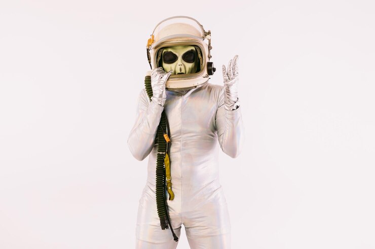Pessoa vestida com um terno prateado e uma máscara alienígena verde, usando um capacete de cosmonauta, sobre fundo branco