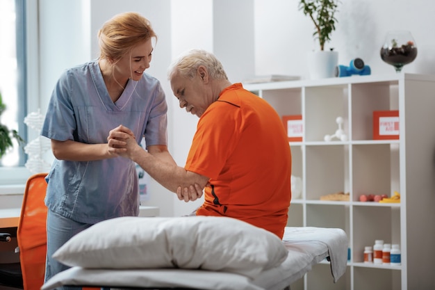Pessoal do hospital. homem idoso sério sentado na cama médica enquanto é ajudado por uma enfermeira