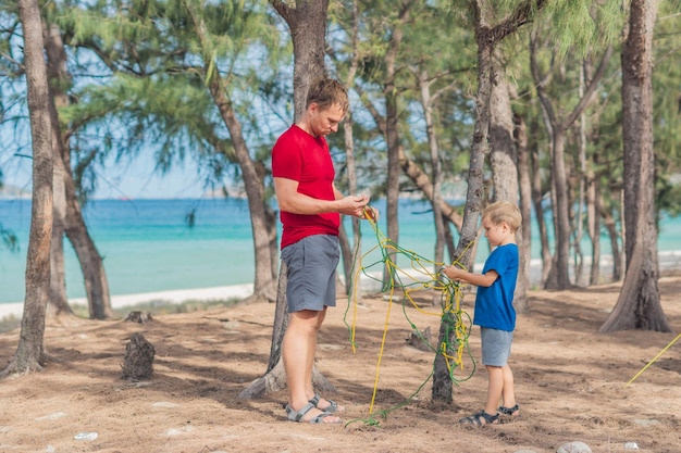 Pessoas acampando turistas de estilo de vida ao ar livre na floresta de verão perto do mar de lazur menino loiro com pai estuda técnicas de sobrevivência praticam métodos de amarrar nós de corda educação natural para crianças