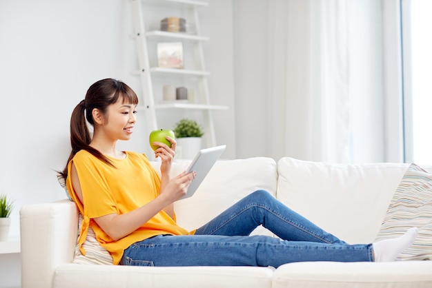 pessoas, alimentação saudável, educação, tecnologia e conceito - feliz jovem mulher asiática sentada no sofá com computador tablet pc e maçã verde em casa