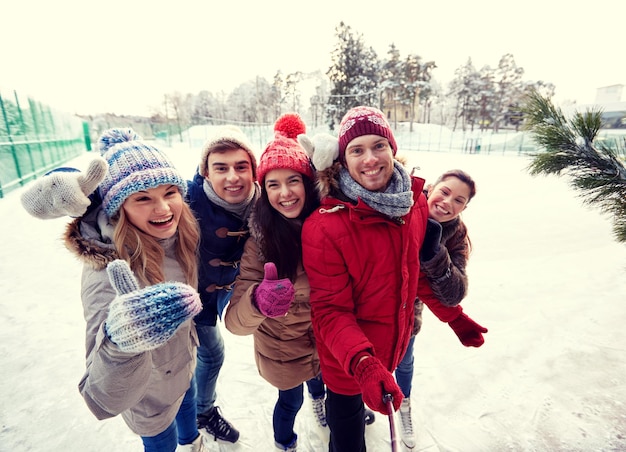 pessoas, amizade, tecnologia e conceito de lazer - amigos felizes tirando foto com smartphone selfie stick e mostrando os polegares na pista de patinação no gelo ao ar livre