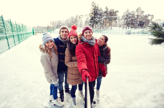 pessoas, amizade, tecnologia e conceito de lazer - amigos felizes tirando foto com smartphone selfie stick na pista de patinação no gelo ao ar livre