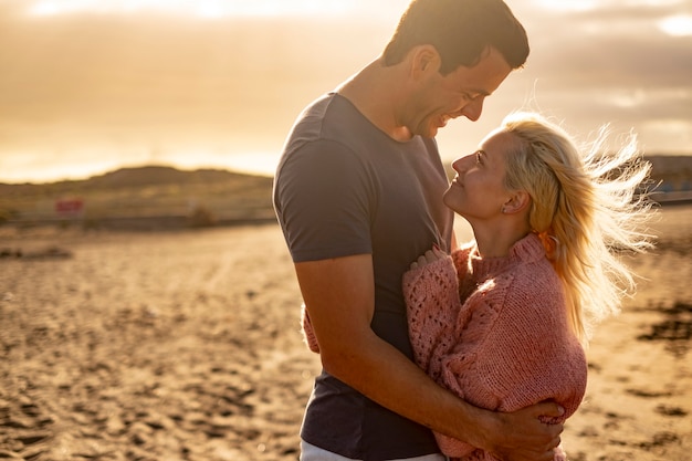 Pessoas apaixonadas e o pôr do sol dourado ao ar livre - casal homem e mulher se abraçam e se olham com a praia ao fundo - conceito de proteção e vida juntos para sempre