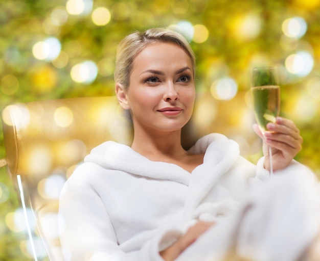 pessoas, beleza, estilo de vida, férias e conceito de relaxamento - mulher jovem e bonita em roupão de banho branco deitado na chaise-longue e bebendo champanhe no spa sobre fundo de luzes de férias