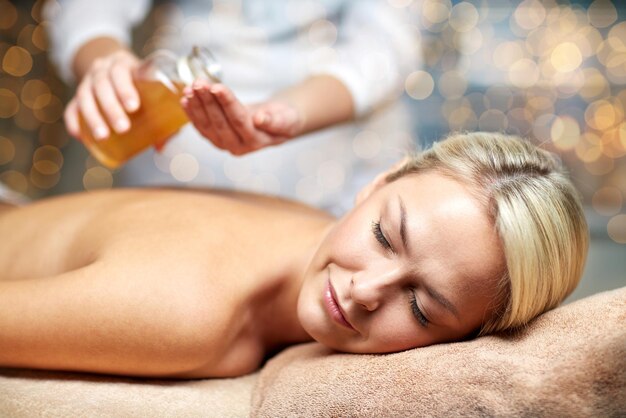 Foto pessoas, beleza, spa, estilo de vida saudável e conceito de relaxamento - close-up de bela jovem deitada com os olhos fechados na mesa de massagem e terapeuta segurando garrafa de óleo no spa