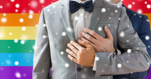 pessoas, celebração, homossexualidade, casamento entre pessoas do mesmo sexo e conceito de amor - close-up de casal gay masculino feliz com alianças de casamento abraçando sobre fundo de bandeira de arco-íris e efeito de neve