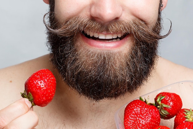 Pessoas comida beleza estilo de vida dia dos namorados conceito closeup retrato jovem segurando um morango e sorrindo Retrato de bonito barbudo e bigode em um fundo cinza