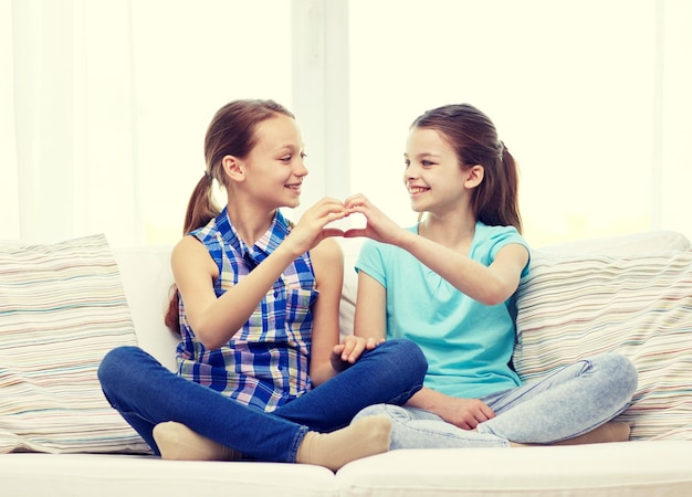 pessoas, crianças, amigos e conceito de amizade - meninas felizes sentadas no sofá e mostrando sinal de mão em forma de coração em casa