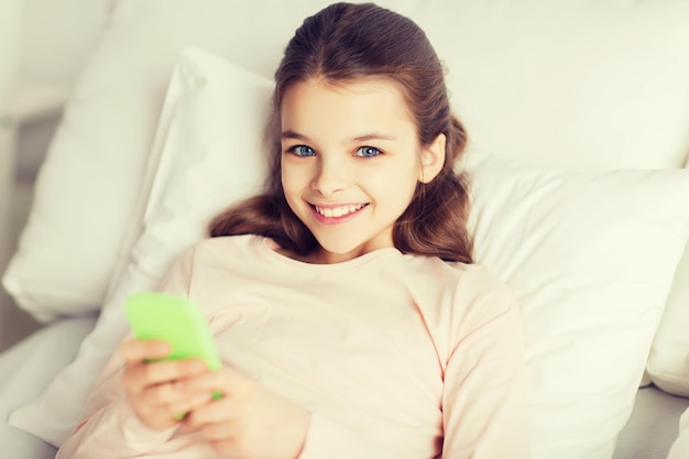 pessoas, crianças, descanso e conceito de tecnologia - garota sorridente feliz acordada com smartphone na cama em casa