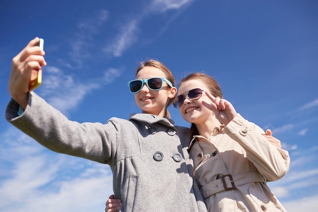 pessoas, crianças, tecnologia, amigos e conceito de amizade - meninas felizes com smartphone tirando selfie ao ar livre