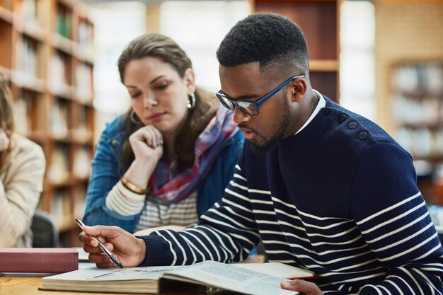 Pessoas da biblioteca e estudando com livros leitura e mesa para exame mulher e homem no campus da faculdade estudantes amigos e trabalho em equipe para informações de teste e mesa com pesquisa e juntos