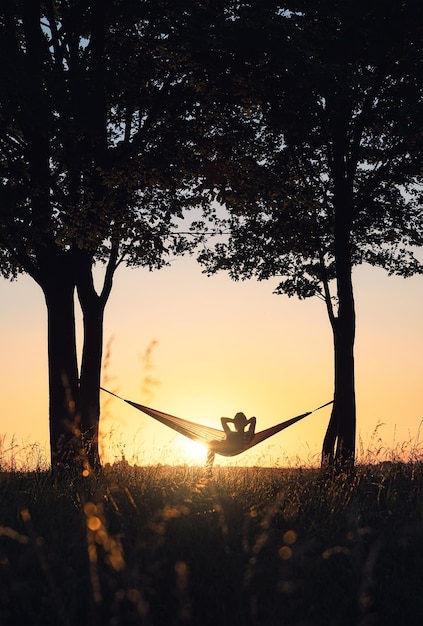 Pessoas de férias A silhueta de uma menina em uma rede entre as árvores Uma rede no fundo do pôr do sol Descanso e relaxamento na natureza