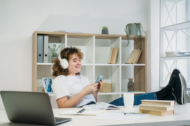 Pessoas de gadgets Lazer dentro de casa Navegação por telefone Comunicação móvel Estudante de mulher de cabelos cacheados com excesso de peso satisfeita em fones de ouvido digitando mensagem no apartamento moderno e leve