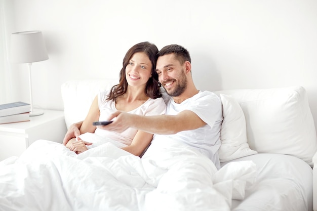 pessoas, descanso, amor, relacionamentos e conceito de televisão - casal feliz com controle remoto deitado na cama em casa e assistindo tv