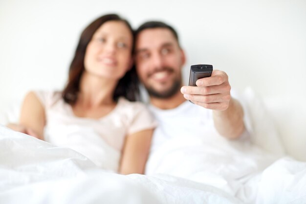 pessoas, descanso, amor, relacionamentos e conceito de televisão - casal feliz com controle remoto deitado na cama em casa e assistindo tv